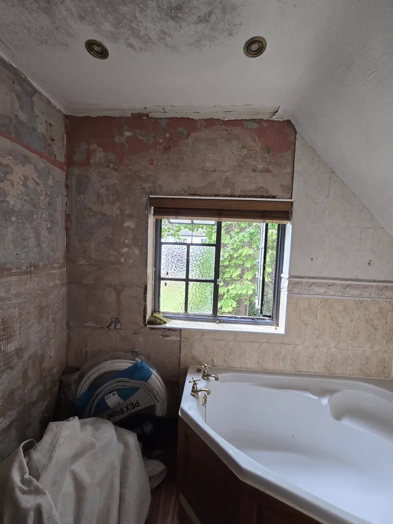 Bathroom mid strip-out with exposed brick wall, a window with a roller blind, and a bath in the corner.
