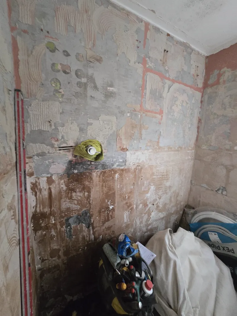 Room corner with patchy plaster repairs on the walls, tools and materials piled on the floor.