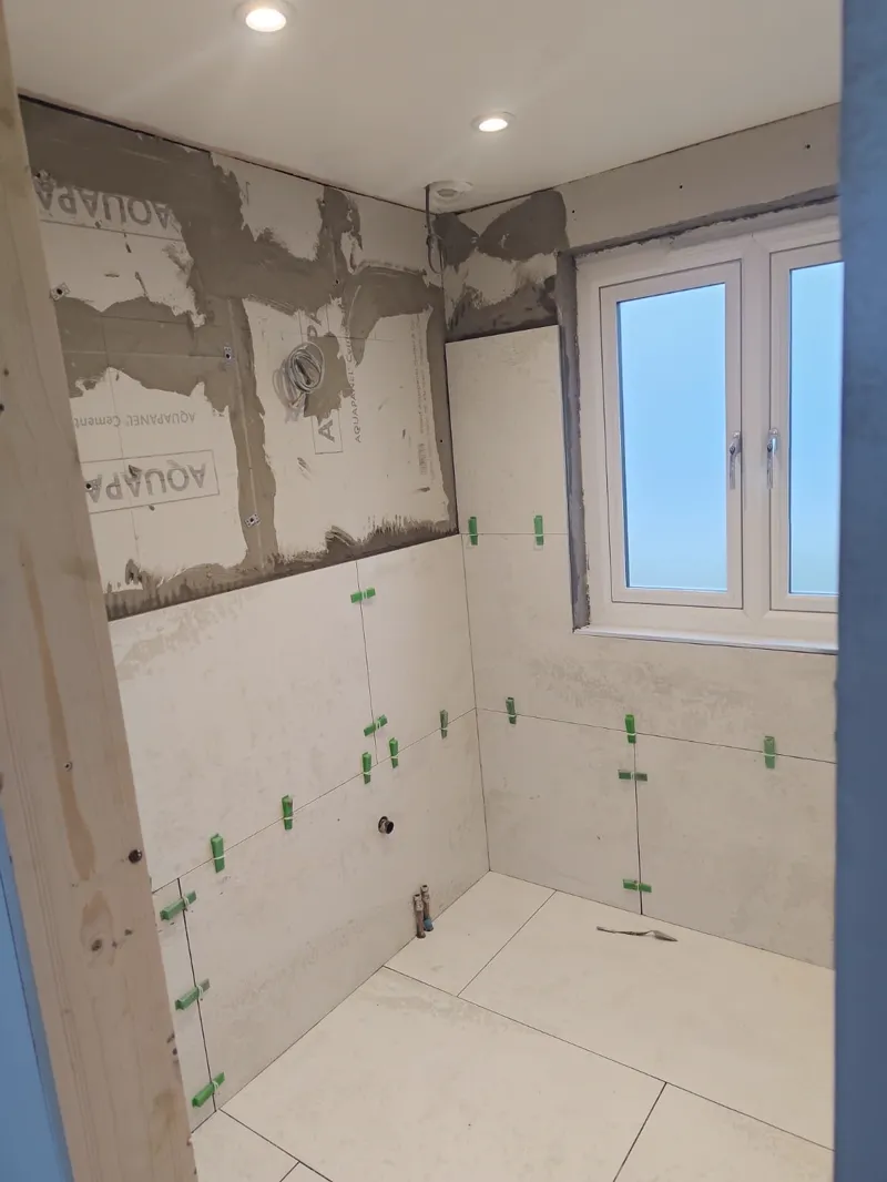 Tiled bathroom corner with downlights fitted, a window, and floor tiles partly laid with spacers.