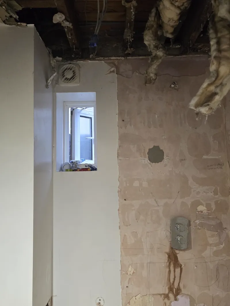 Bathroom mid strip-out with a small window and extractor fan, exposed block wall and ceiling joists above.
