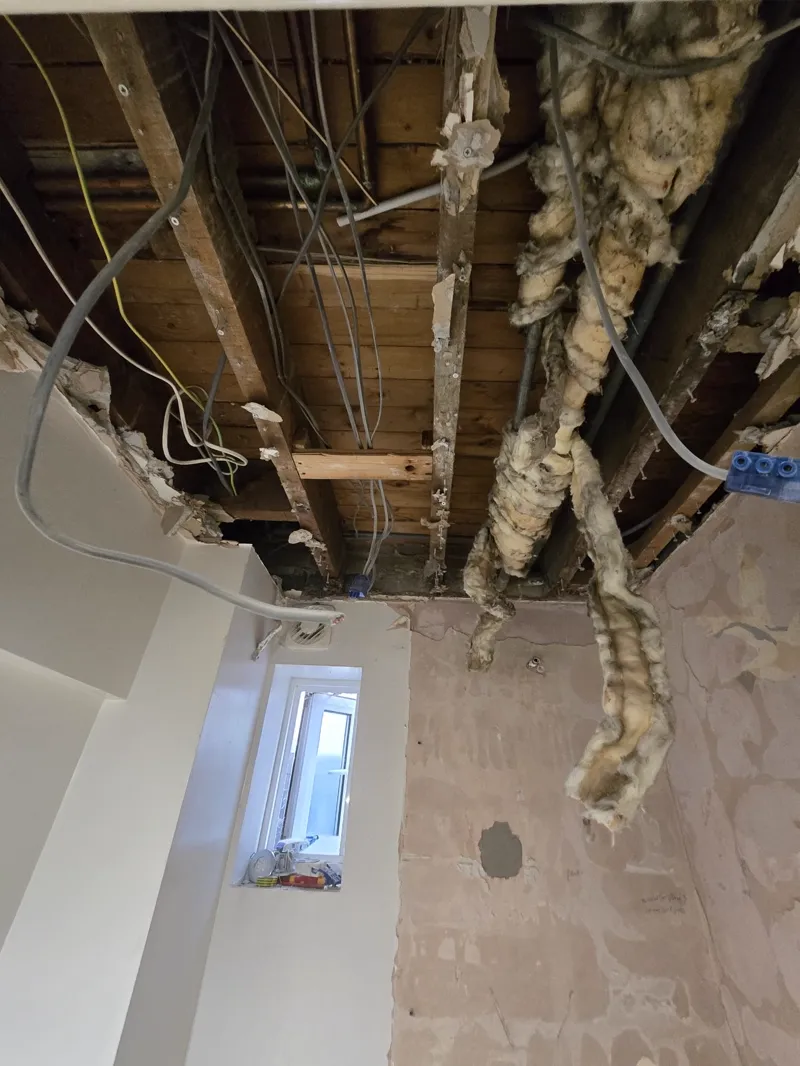 Ceiling joists, wiring and loft insulation exposed above a stripped-out bathroom, small window visible.