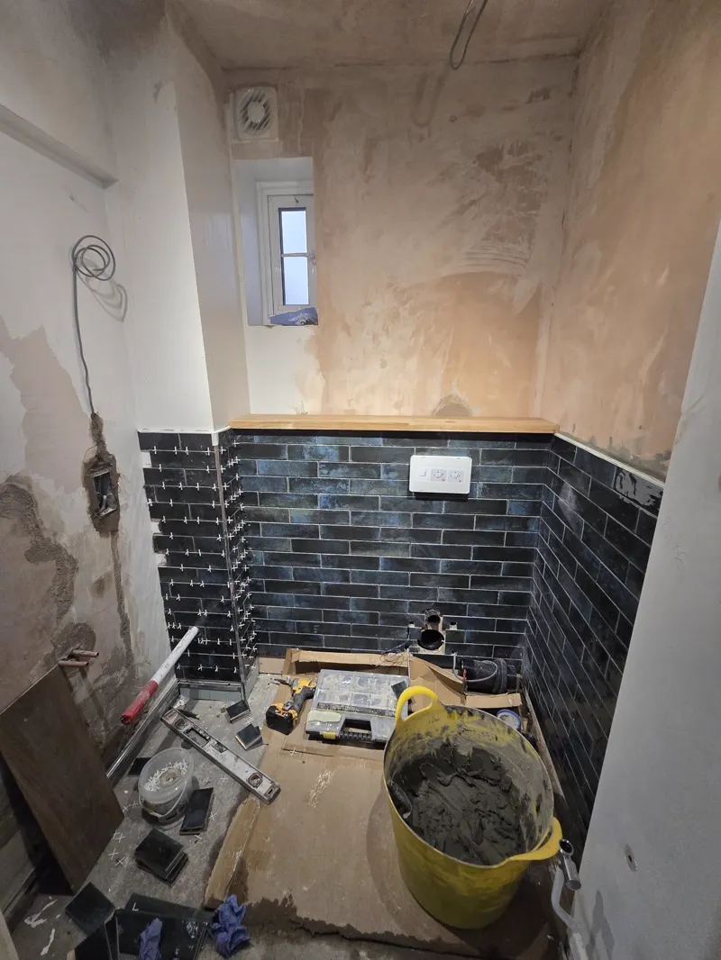 Bathroom mid-tiling with black metro tiles partway up the wall, damp-stained plaster above, tools and a cement bucket on the floor.