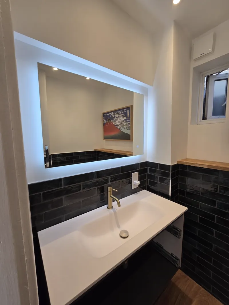 A backlit vanity mirror over a black metro-tiled wall and a countertop basin with a mixer tap.